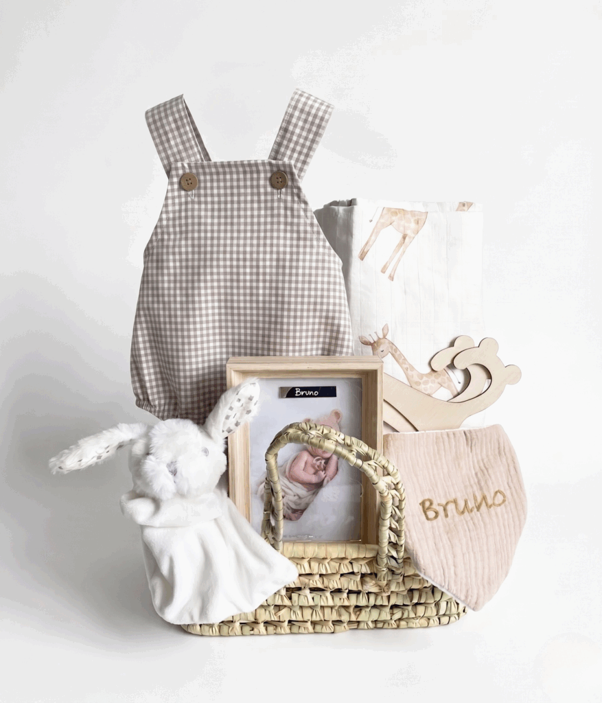 Cesta de mimbre con pelele de cuadros beige, manta de jirafas, peluche de conejo doudou y accesorios personalizados con el nombre bordado.