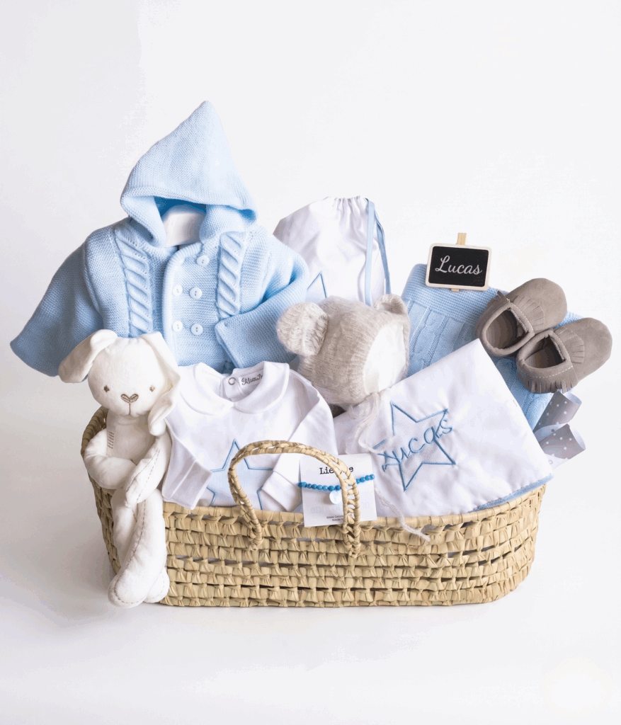 Canastilla de bebé azul en capazo con chaqueta de punto, patucos, peluche de conejo y manta bordada con el nombre.