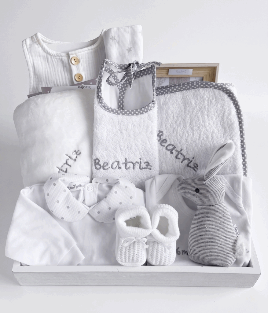 Canastilla de regalo para bebé personalizada con nombre bordado, que incluye ropa blanca, babero de toalla, patucos de punto y peluche de conejo gris.